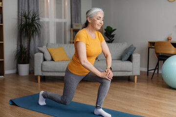 Positive sporty senior woman keeping fit at home, doing lunges, exercising on yoga mat in living...