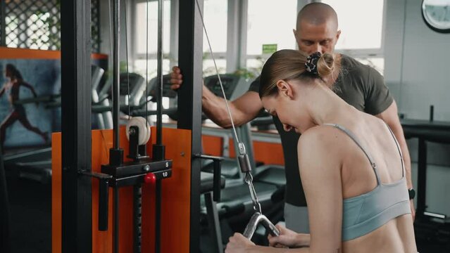 Caucasian blond athletic woman in her 20s with her hair tied up in a bun, focusing on doing an exercise for her triceps and back muscles. Personal male trainer in the background. High quality 4k