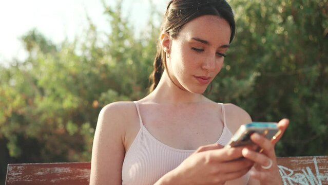 Young athletic woman with braided pigtail wearing beige sports top is sitting on bench with mobile phone in her hands, watching social networks, photos and videos