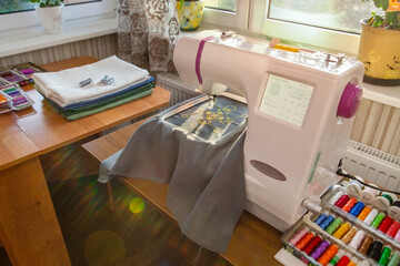 Home sewing workshop, workplace opposite the window, with a modern embroidery machine that creates...