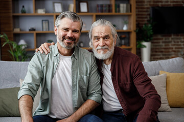 Handsome smiling adult man hugging senior mature father, looking at camera. Happiness, love and care. Two generations family close up portrait. Fathers day concept