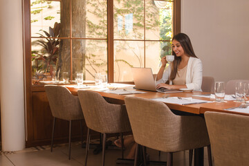 Young smiling business girl drinking coffee while using her laptop in the restaurant.