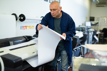 Man working in publishing facility, loading large format paper in printer. - obrazy, fototapety, plakaty