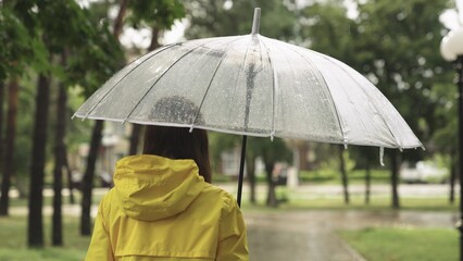 a young girl with a transparent umbrella walks in the rain. woman in a yellow raincoat. view from the back. autumn summer spring walk park. Slow motion. wet drops rain drip transparent umbrella.
