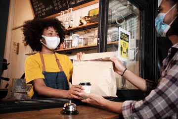 African American female barista with face mask works in the cafe with social distance, takeaway coffee for a customer, new normal service of small business coffee shop in COVID19 quarantine lifestyle.