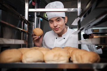 Young Asian male chef in white cook uniform and hat showing tray of fresh tasty bread with a smile,...