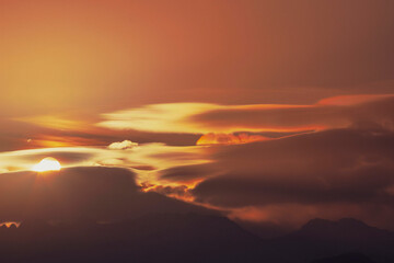 The sun rises among the clouds above the silhouettes of mountain hills.