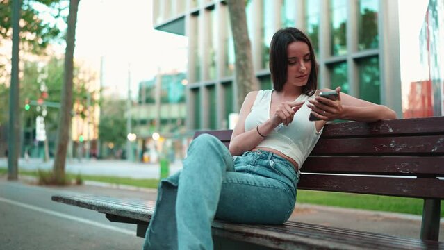 Beautiful woman with freckles and dark loose hair wearing white top sits on bench with phone in her hands. Cute girl browsing messages, photos and videos on mobile phone 