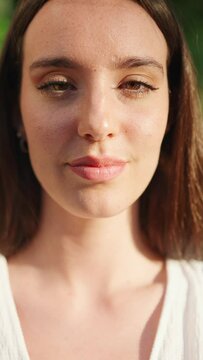 VERTICAL VIDEO: Young woman with freckles and dark loose hair wearing white top and jeans opens her eyes and smiles. Beautiful girl with long eyelashes smiling broadly at the camera