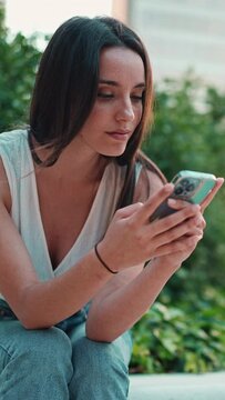 VERTICAL VIDEO: Cute young woman with freckles and dark loose hair wearing white top is using mobile phone. Beautiful girl sits in city park looking through photos, videos on mobile phone