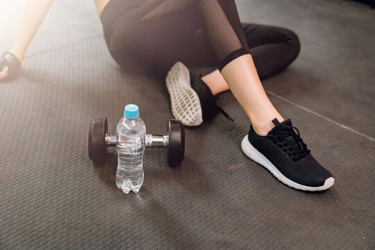 Woman sitting shoelaces with protein shake and dumbbell at fitness gym after running exercise workout for cardio and muscle building.