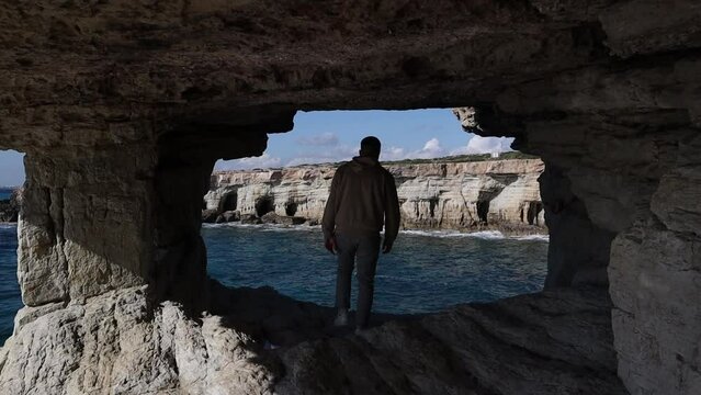 A man walks and sits in the hole in a cave with a beautiful view of the sea near cape Greco in a national park with turquoise water sea in Ayia Napa, Cyprus. 