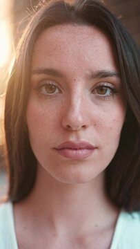 VERTICAL VIDEO: Close-up of young woman with freckles and dark loose hair and long eyelashes wearing white top looking straight at the camera. Beautiful girl on modern city background