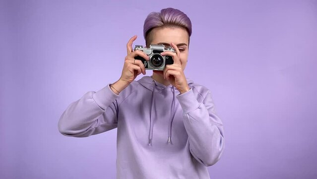 Cheerful young adult woman with trendy look taking pictures with old retro film camera, turning advance lever photographing with joy isolated on purple background indoors. Charming girl wearing hoodie