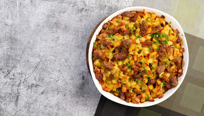 Oven baked meat with potatoes and herbs in a white baking dish on a dark grey background. Top view, flat lay