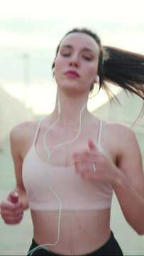 VERTICAL VIDEO: Young athletic woman with long ponytail wearing beige sports top in wired headphones, runs along the pedestrian crossing to the beach