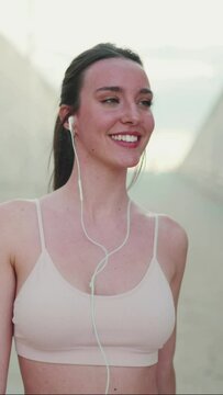 VERTICAL VIDEO: Young athletic woman with long ponytail wearing beige sports top in wired headphones, walking dancing and smiling
