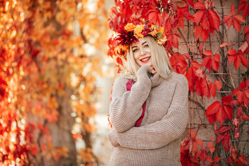 Smiling blonde in knitted warm sweater and bright wreath of fallen leaves stands near wall , braided with red girlish grapes in autumn in park. Beautiful autumn. Seasons. Indian summer.
