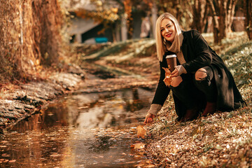 Stylish blonde crouching near stream pulls her hand to water in city park. She is illuminated by...
