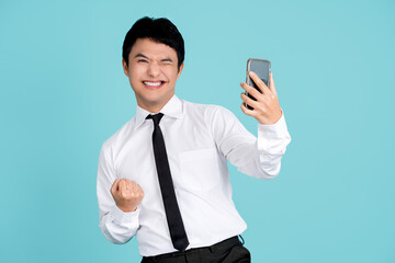 Cheerful handsome asian man holding smartphone with fists clenched celebrating victory expressing success on light blue background.