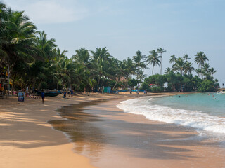 Hikkaduwa, Sri Lanka - March 4, 2022: Beautiful view of the beach in Hikkaduwa with bright green palm trees. People relax and swim in the Indian Ocean