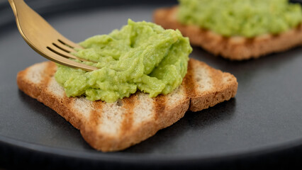 Preparing avocado toast. Hands with fork spreading mashed avocado on toasted whole  bread. Vegan food concept