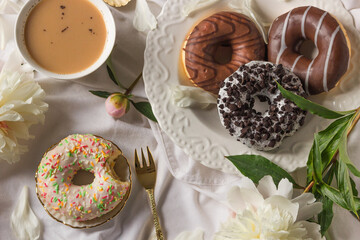 Top view of breakfast in bed with various delicious glazed donuts on the plate, cup of coffee and fresh peony flowers.