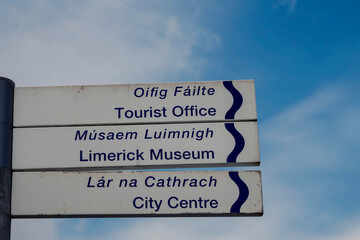 Sign in English and Irish language in Limerick city with direction to Tourist office, Museum and city centre. Blue cloudy sky background.