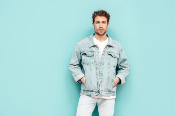 Portrait of handsome confident stylish hipster lambersexual model with curly hairstyle. Sexy man dressed in jeans jacket. Fashion male isolated on blue wall in studio