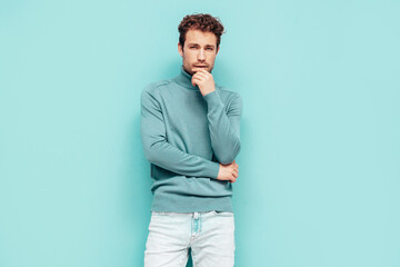 Portrait of handsome confident  model. Sexy stylish man dressed in  sweater and jeans. Fashion hipster male with curly hairstyle posing near blue wall in studio. Isolated