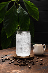 Espresso tonic in the making. A highball glass filled with ice cubes, tonic soda water, coffee beans, milk jug, wooden background