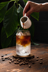 Espresso tonic in the making. Adding coffee into a highball glass filled with ice cubes and tonic soda water, coffee beans, milk jug, wooden background - obrazy, fototapety, plakaty