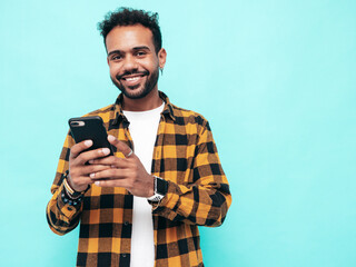 Handsome smiling hipster model. Sexy unshaven man dressed in yellow  summer shirt and jeans clothes. Fashion male posing near blue wall. Holding smartphone. Looking at cellphone screen. Using apps