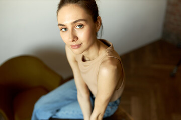 Upper angle image with selective focus on beautiful face of adorable and charming Caucasian female sitting on table in office or classroom. Student posing in jeans and tank top during break