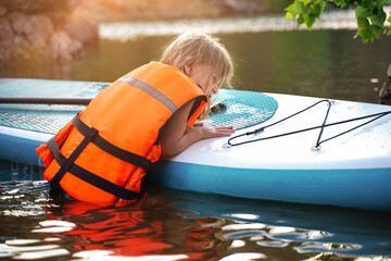 European child girl in a life jacket climbs on a SUP board floating on the lake