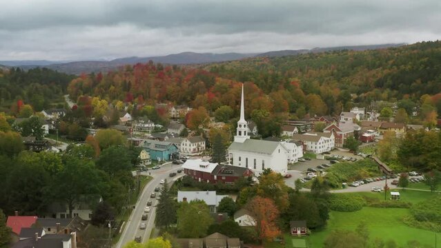 Church building in small town with scenic fall foliage forest landscape background on cloudy autumn day. East Coast Vermont USA. Cinematic 4K aerial of white wooden old historic Stowe community church