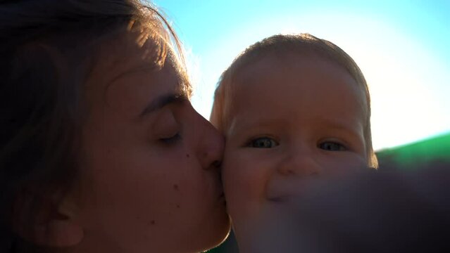 Caring mother kisses cheek of little son standing in yard on sunny day. Portrait of happy woman with toddler on blurred background closeup slow motion