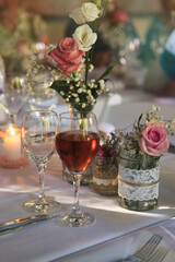 Table decoration arrangement of glasses, flowers, cutlery, napkins, small burning candles, and a full glass of red wine ready to start the wedding dinner, with guests sitting in the blurry background.