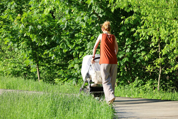 Slim woman walking with baby pram in green park. Young mother, leisure at summer nature