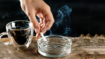 Glass ashtray, coffee and lit cigarette in hand on black background wooden table