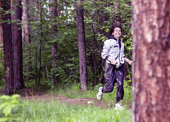 Young woman running in the forest along the path between green trees. Happy smiling female face while jogging