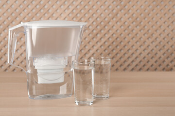 Filter jug and glasses with purified water on wooden table indoors. Space for text
