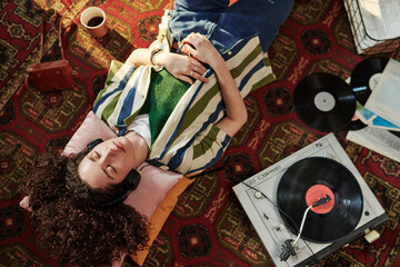 Above angle of young female in casualwear enjoying music in headphones while resting on red carpet on the floor