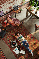 Overview of young woman in stylish casualwear resting on the floor while enjoying music on vynil...
