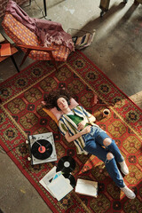 Girl in casualwear lying next to record player with vynil disks on red carpet in living room and...