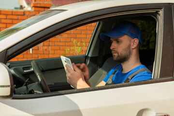 Naklejka premium Bearded man in blue cap sits on driver seat and makes notes about internal car details on tablet. Auto mechanic types on tablet diagnosing car state closeup