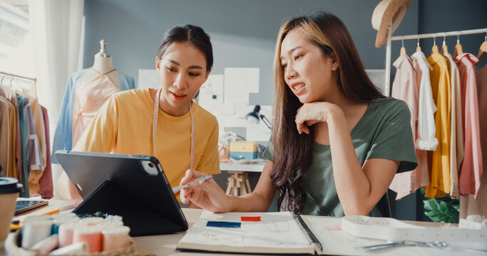 Two professional Asia cheerful lady fashion designer teamwork with casual site on table use tablet meeting discuss final design share idea in shop. Lady tailor partner start up business cloth shop.