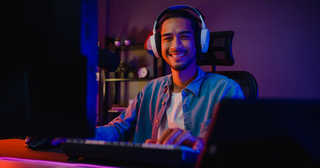 Happy asia guy gamer wear headphone participation play video game colorful neon lights computer and smiling look at camera in living room at night modern house. Esport streaming game online. © tirachard