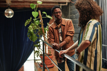 Young intercultural couple in stylish casual attire standing on balcony with green domestic plant...