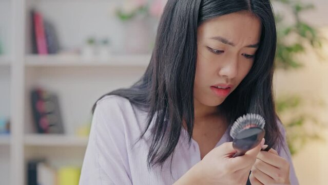 Asian woman combing weak hair and looking at brush, displeased with split ends